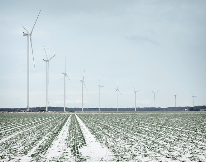 Tuuliturbiineja pellolla vierekkäin, maassa hento lumikerros jonka läpi näkyy vielä vihreää maata.