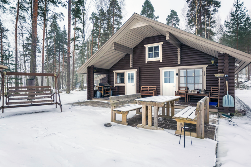 Kuva tummanruskeasta mökistä, pihan edustalla puiset pöytä ja penkit sekä pihakeinu, ympäröivä metsä on talvinen