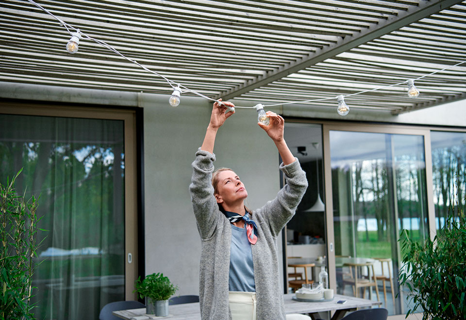 Nainen ripustaa talon pihalla kirkkaita juhlavaloja terassin katon alle