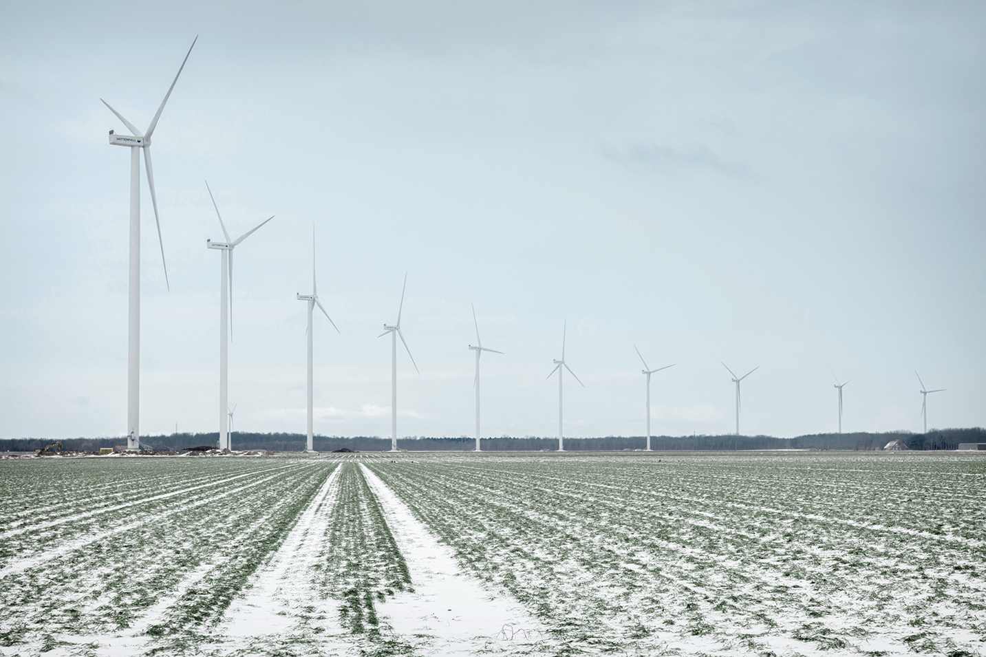 Tuuliturbiineja pellolla vierekkäin, maassa hento lumikerros jonka läpi näkyy vielä vihreää maata.