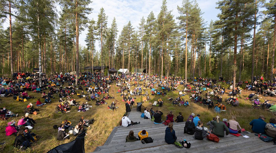 Laajakuva suuresta ihmismäärästä metsäaukealla, osa istuu puisilla portailla ja loput maassa