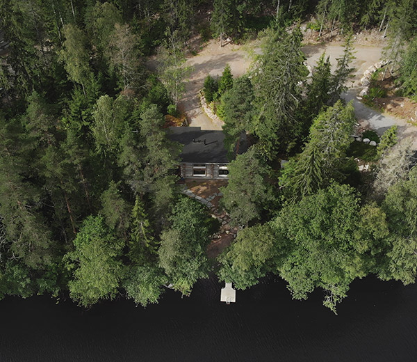 Ilmakuva jossa tuuhean kuusimetsän reunalla puumökki sekä tie