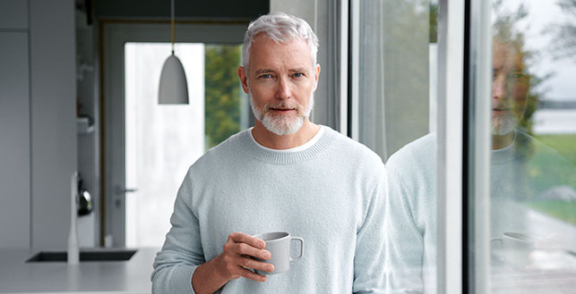 Vanha mies seisoo ikkunan vieressä kahvikuppi kädessä