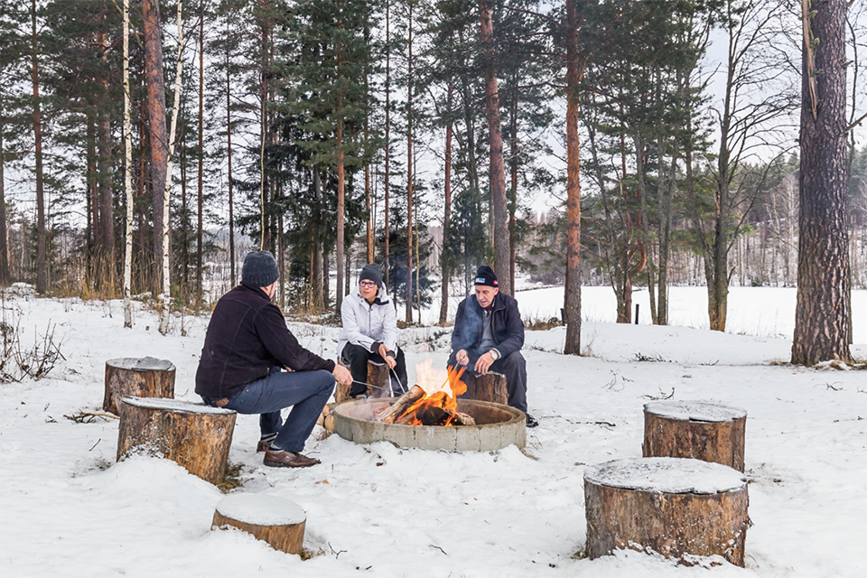 Kolme henkilöä istuu nuotion äärellä talvimaisemissa kantojen päällä ja paistaa makkaraa tikuissa.