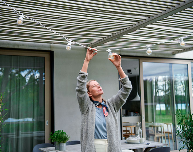 Nainen ripustaa talon pihalla kirkkaita juhlavaloja terassin katon alle