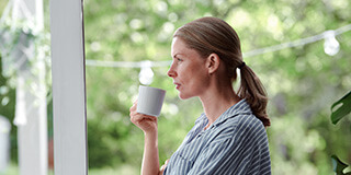 Nainen pitää kädessä kahvikuppia ja katsoo ulos ikkunasta. 