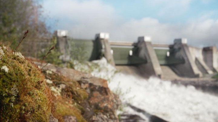 Kuva: etualalla sammalainen kivi, taustalla vesivoimalaitos