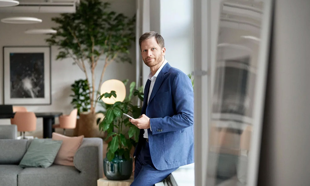 Mies sinisessä puvussa nojaa ikkunalautaan ja katsoo kameraan